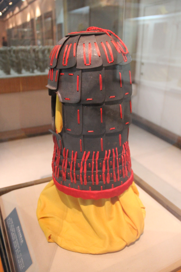 Ein Helm in einer Glasvitrine mit einem gelben Tuch darüber, begleitet von einer Texttafel auf der linken Seite und Deckenlampen im Hintergrund.