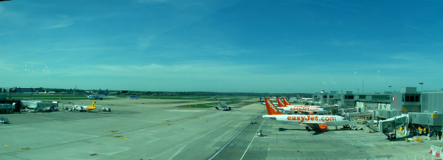 Eine Flughafenszene mit Flugzeugen, Gebäuden, Pfählen, Fahrzeugen, Bäumen und einem Himmel darüber, einer Straße darunter und einem Zaun im Vordergrund.