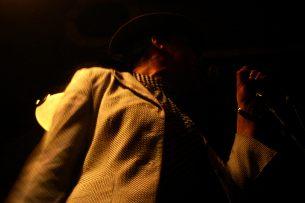 Ein Mann mit Hut und Jacke hält ein Mikrofon, das vor einem unscharfen Hintergrund hervorgehoben ist.