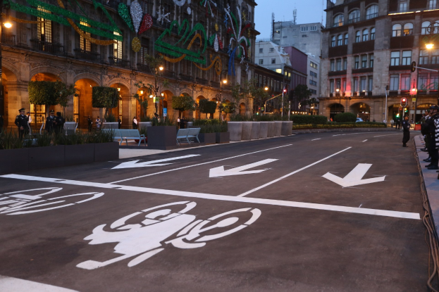 Eine Stadtstraße mit einem Radweg, Gebäude, Straßenlaternen, Verkehrspoller, Verkehrsampeln, Menschen auf dem Gehweg, Bänke, Pflanzen, Bäume, dekorative Gegenstände und einen klaren blauen Himmel.