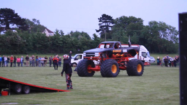 Ein Monstertruck steht auf einer Rampe mit einem Van und Autos in der Nähe, umgeben von Gruppen von Menschen, Bäumen, Gras, einem Gebäude und einem klaren Himmel.