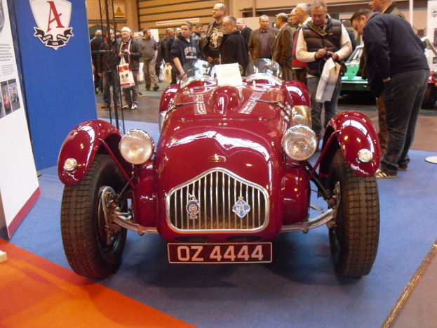 Ein Fahrzeug auf einer Ausstellung mit zwei Schildern auf der linken Seite, umgeben von Menschen und anderen Fahrzeugen, mit einer Wand im Hintergrund.