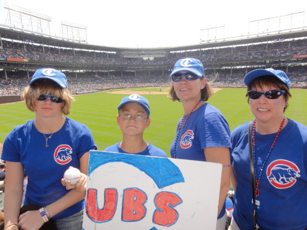 Drei Frauen in Kappen und Brillen, ein Junge in Brille und Kappe mit einem Ball und einer Tafel, die in einem Stadion mit Zuschauern und Feld hinter ihnen und einem Himmel mit wenigen Lichtern steht.