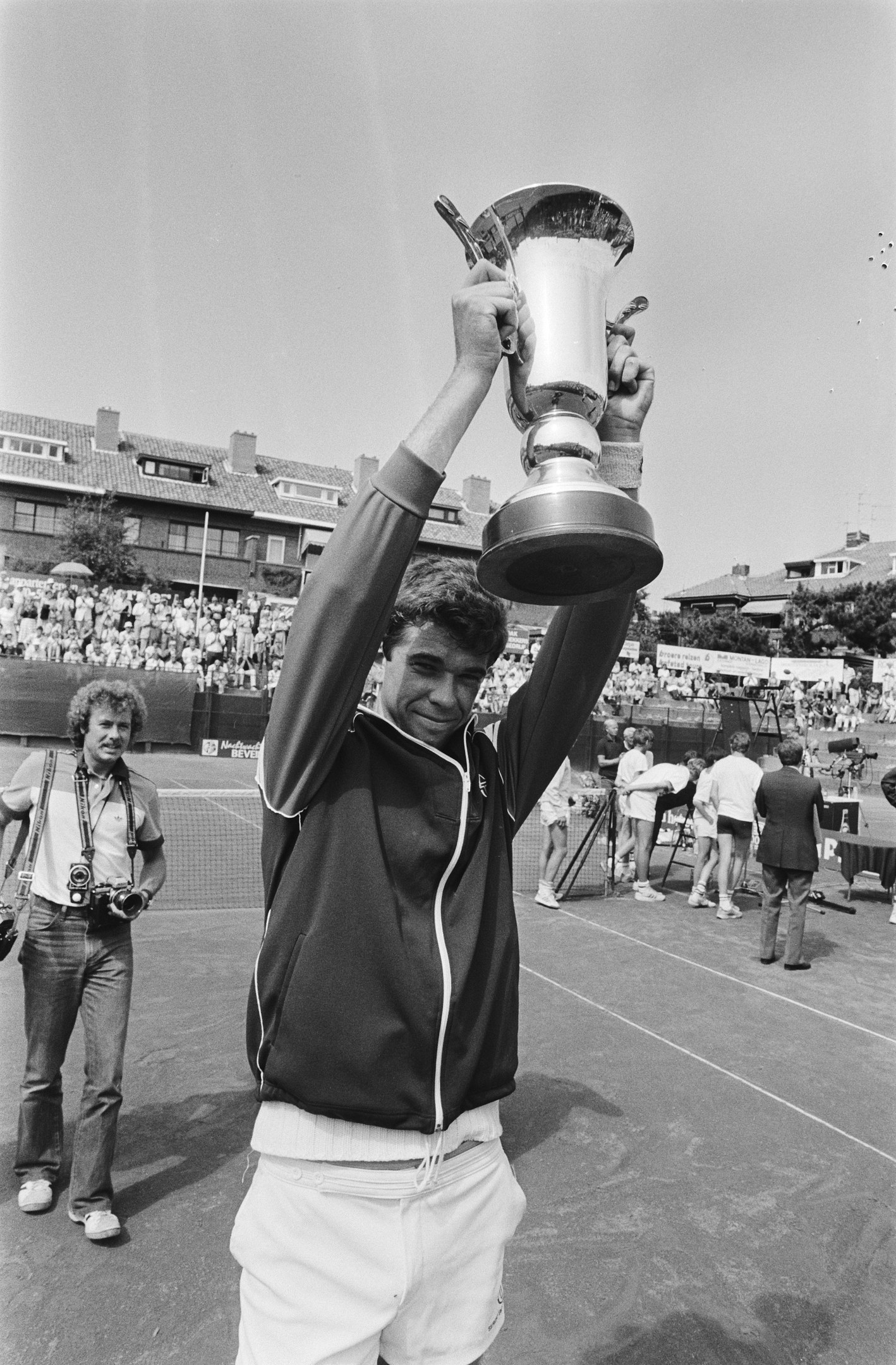 Ein Mann hält triumphierend einen Pokal auf einem Tennisplatz hoch, umgeben von einer Menge mit Kameras, mit Gebäuden, Bäumen und einem klaren blauen Himmel im Hintergrund.