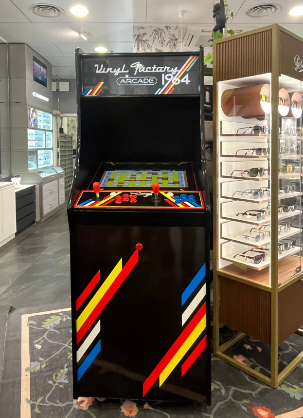 Ein Geschäft mit einem zentralen Retro-Arcade-Automaten, einer rechten Glasvitrine und Hintergrundschränken, Fernseher und Beleuchtung im Retro-Stil.