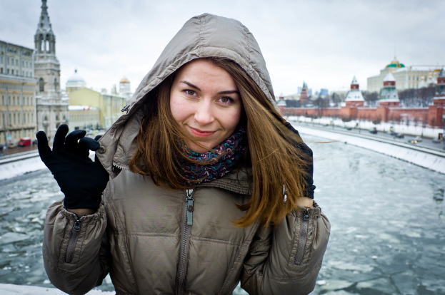 Eine Frau in einer Jacke steht vor einer zugefrorenen Wasserfläche, mit Gebäuden und fahrenden Fahrzeugen auf beiden Seiten und einem Himmel im Hintergrund.