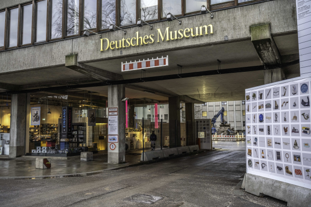 Außenansicht des Deutschen Museums in Berlin, ein großes Gebäude mit Glasfenstern, Säulen und einem Namensschild, neben einer informativen Anzeigetafel, mit anderen Gebäuden, Bäumen und Himmel im Hintergrund.