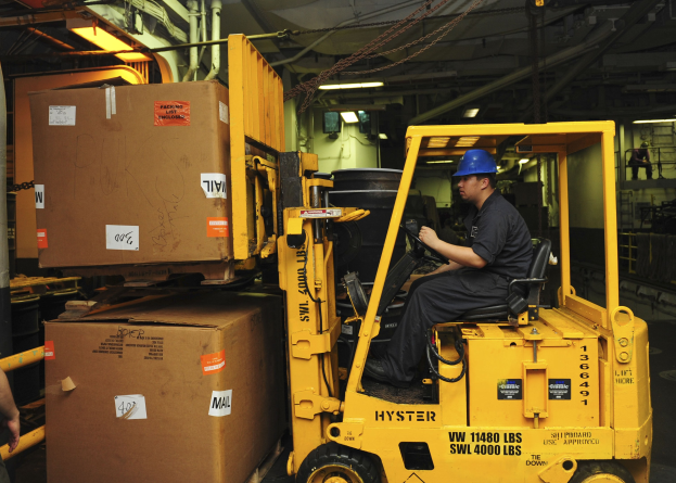 Ein Mann in einem schwarzen Kleid und einem blauen Helm bedient einen gelben Gabelstapler in einem Lagerhaus, mit Kartons zu seiner Linken und industriellen Elementen wie Rohren und Lampen im Hintergrund.