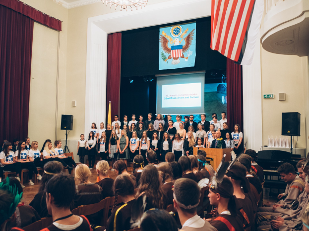 Eine Gruppe von Menschen steht vor einem sitzenden Publikum auf einem Podium mit Mikrofon, flankiert von Fahnen und Lautsprechern, während eines 2. Wochen Kunst- und Kultur-Events.