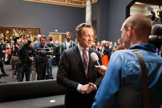 Ein Mann im Anzug und Krawatte spricht mit einem Reporter und hält ein Mikrofon in der Hand, im Hintergrund ist eine Menschenmenge zu sehen, einige halten Kameras, im Vordergrund steht ein Sofa und an der Wand ist ein Foto Rahmen und Lampen zu sehen.