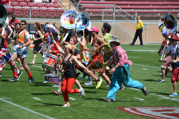 Eine Gruppe von Menschen versammelt sich im Freien mit Musikdrums, verschiedenen Instrumenten, Stühlen, Treppen und einem Zaun im Hintergrund.