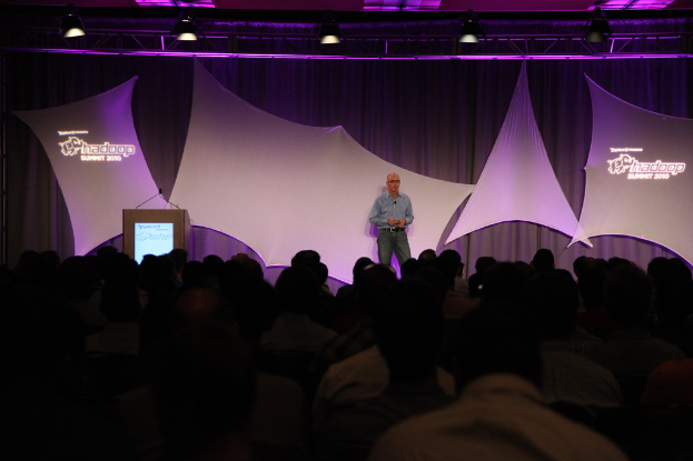 Ein Mann in einem blauen Hemd steht auf einer Bühne mit schwarzen und weißen Vorhängen und spricht zu einem sitzenden Publikum unter einem Metallrahmen mit Scheinwerfer.
