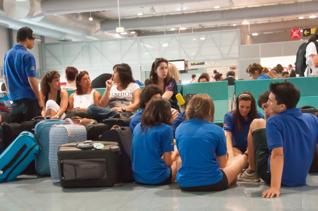 Ein geschäftiger Flughafeninnenraum mit vielen Menschen, die auf Flüge warten, Gepäck auf der linken Seite und Glastüren mit Richtungsschildern im Hintergrund.