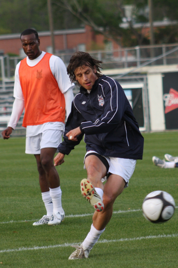 Zwei Fußballspieler auf einem Feld, einer in einer schwarzen Jacke und weißen Shorts, die einen Ball treten, der andere in einem orangen Jackett dahinter, mit einem Zaun und Bäumen im Hintergrund.