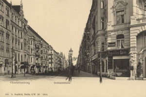Schwarzes und weißes Foto einer Stadtstraße in Charlottenburg, Deutschland, mit Gebäuden, Straßenlaternen, Lampen, Namensschildern, Fahrzeugen, Fußgängern und Gittern unter einem klaren Himmel.