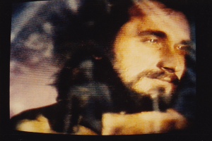 Ein Mann mit langem dunklem Bart und Haar, der ein weißes Hemd trägt, ist auf einem Fernseher mit dunklem Hintergrund zu sehen und schaut direkt in die Kamera.