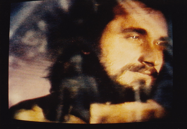 Ein Mann mit langem dunklem Bart und Haar, der ein weißes Hemd trägt, ist auf einem Fernseher mit dunklem Hintergrund zu sehen und schaut direkt in die Kamera.