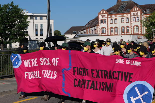 Eine Gruppe von Menschen mit Masken und einem pinken Banner mit der Aufschrift "Wir haben genug von fossilen Brennstoffen, bekämpft den extraktiven Kapitalismus" steht auf einer Straße, einige halten Schirme, mit Gebäuden, Bäumen, einem Pfahl und einem klaren blauen Himmel im Hintergrund.
