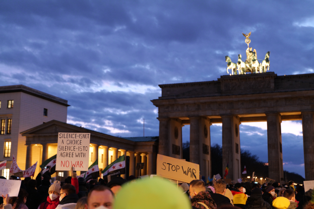 Eine große Gruppe von Menschen mit Schildern und Mützen vor dem Brandenburger Tor in Berlin, Deutschland, mit Fenstern, Säulen und Statuen des Tors, sowie Bäumen und einem bewölkten Himmel im Hintergrund.