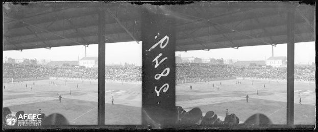 Ein Schwarz-Weiß-Foto eines Fußballspiels in einem Stadion mit Spielern auf dem Boden und Zuschauern in den Rängen, umgeben von Gebäuden, Bäumen und einem klaren Himmel, mit dem Text "Piazza della Repubblica, Rom, Italien" in der linken unteren Ecke.
