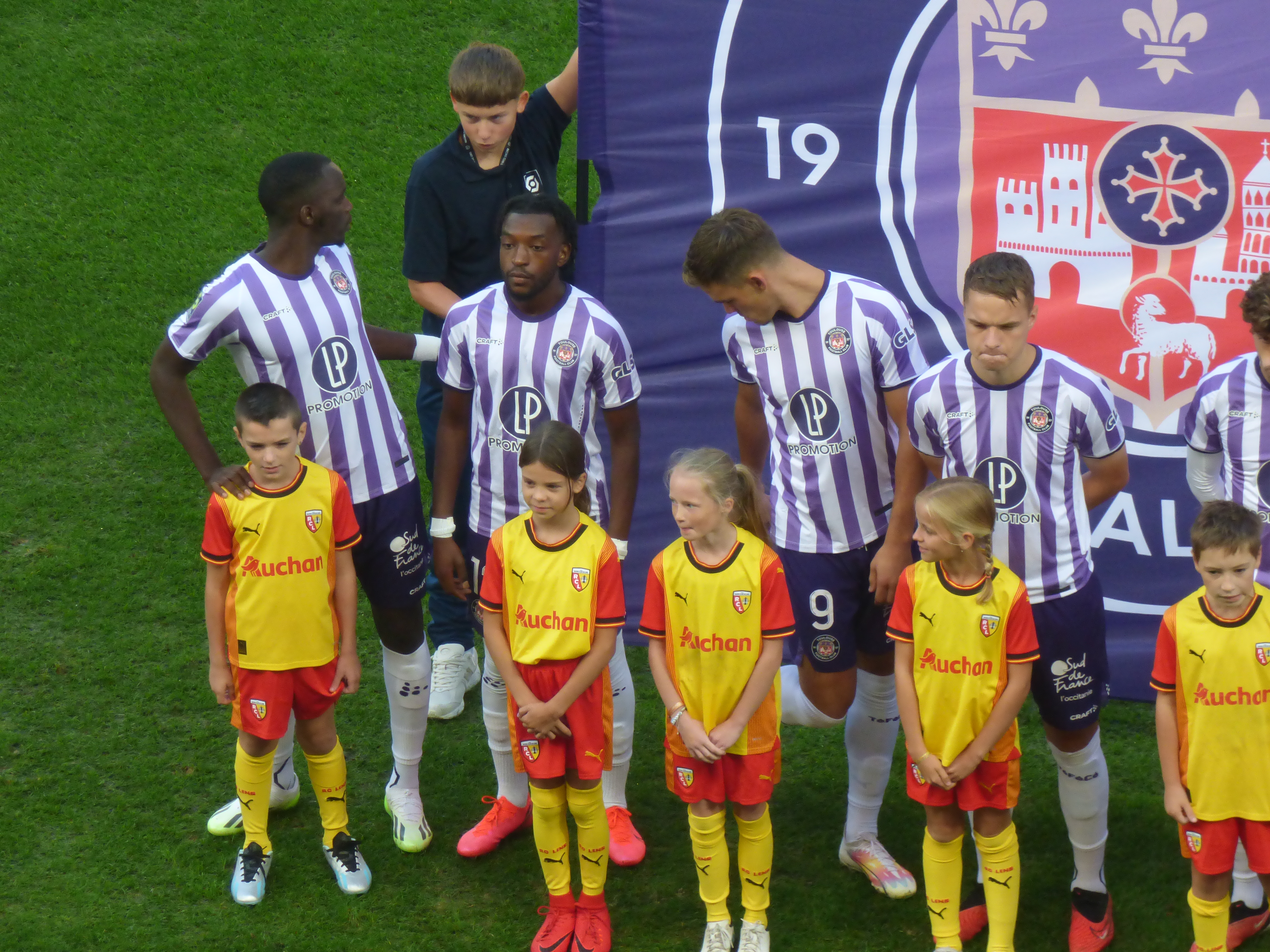 Eine Gruppe junger Menschen auf einem saftig grünen Fußballfeld, mit einem Banner im Hintergrund, auf dem "Ligue 1" steht, einige tragen Trikots und andere kurze Hosen, unter einem strahlend blauen Himmel.