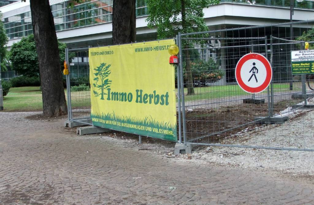 Ein Zaun mit einem Banner, Bäumen und Pflanzen links, einem Gebäude im Hintergrund und einem Kopfsteinpfad unten.