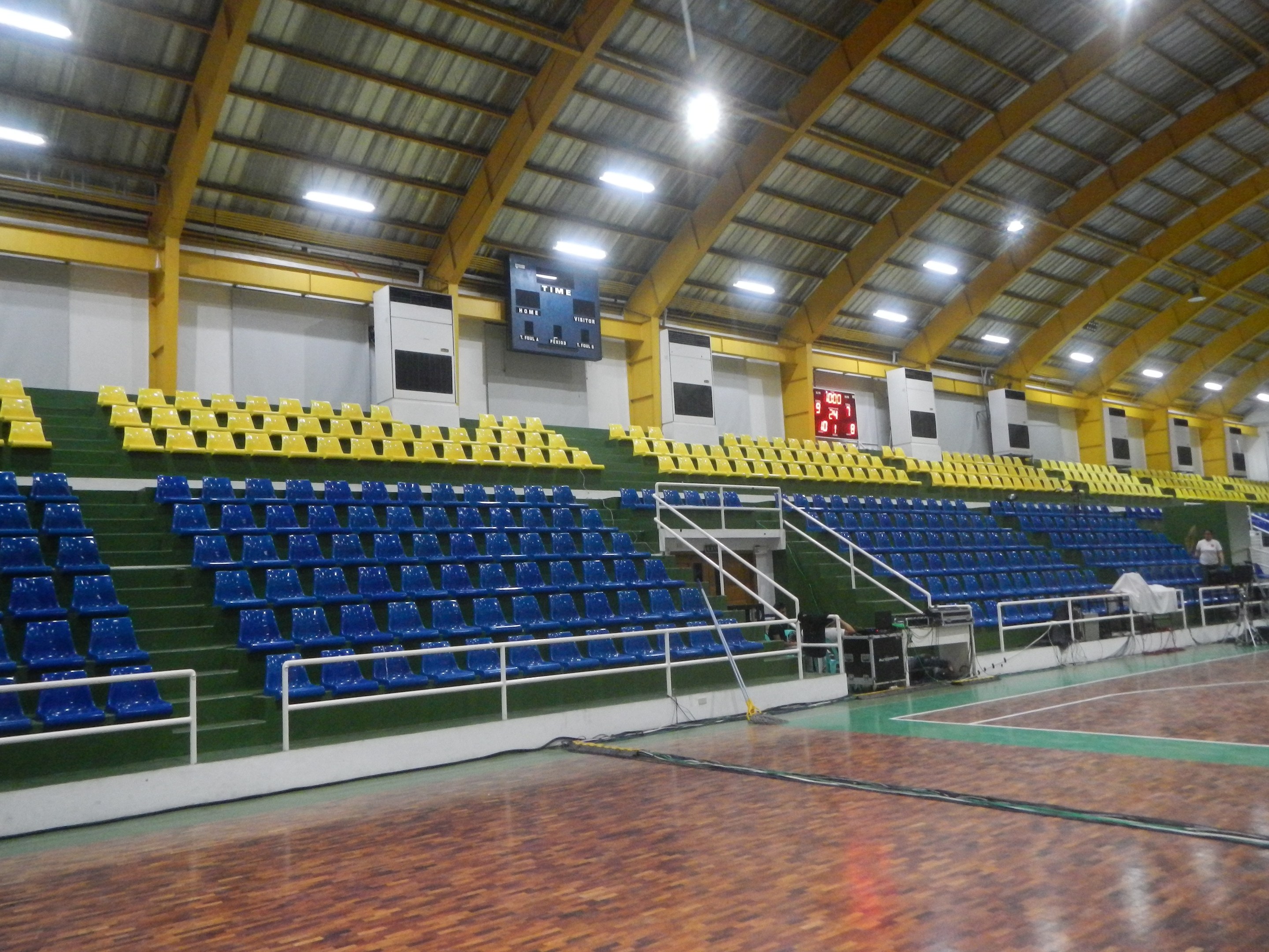 Großer Hallensportplatz mit Holzboden, blauen und gelben Sitzplätzen, Geländern, Treppen, wandmontierten Bildschirmen, Deckenleuchten und einigen Menschen.