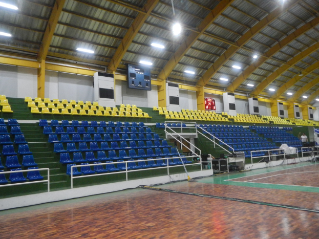 Großer Hallensportplatz mit Holzboden, blauen und gelben Sitzplätzen, Geländern, Treppen, wandmontierten Bildschirmen, Deckenleuchten und einigen Menschen.