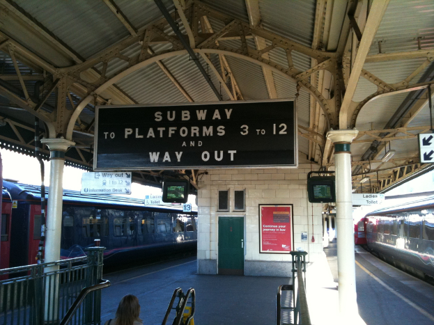 Zwei Züge stehen neben einem Bahnsteig mit Bildschirmen und Tafeln an der Decke, eine Person ist in der Nähe der Treppe und eine Tür ist an der Wand.