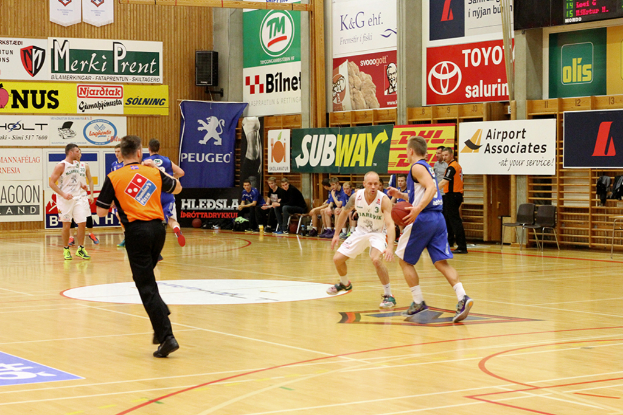 Ein Basketballfeld mit Spielern im Vordergrund und einer holzget├Ąfelten Wand mit Postern und Werbetafeln im Hintergrund.