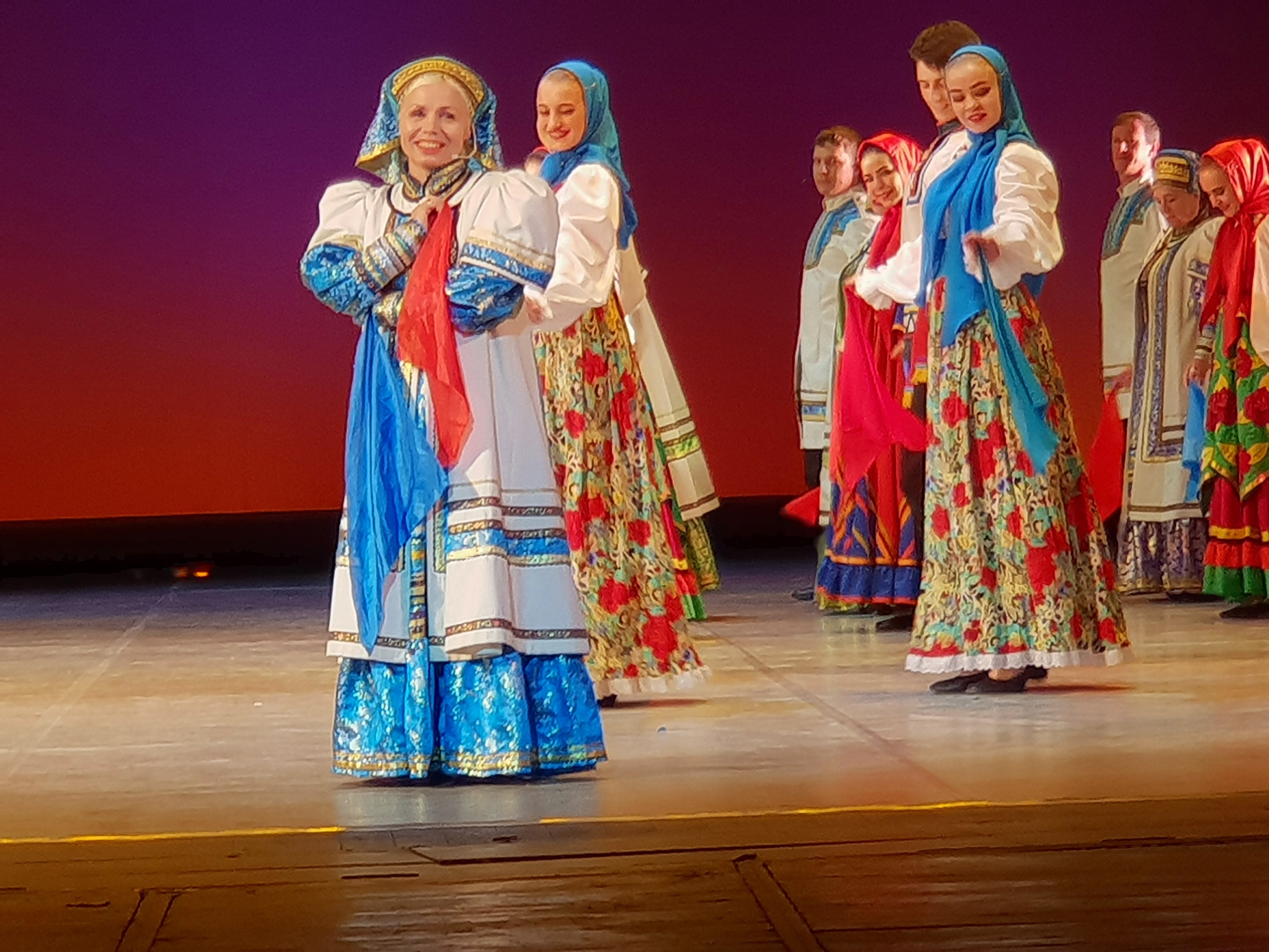 Eine Gruppe von Menschen in farbenfrohen, detaillierten traditionellen ukrainischen Kostümen, die auf einer Bühne lächeln.