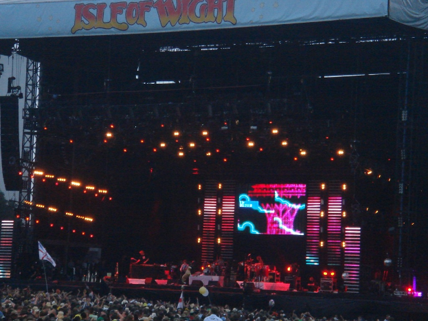 Eine Bühne mit Lichtern, ein Bildschirm und Musiker, die Instrumente spielen, mit einem Banner oben, einer Menge unten und Fahnen mit Stangen und Ballons.