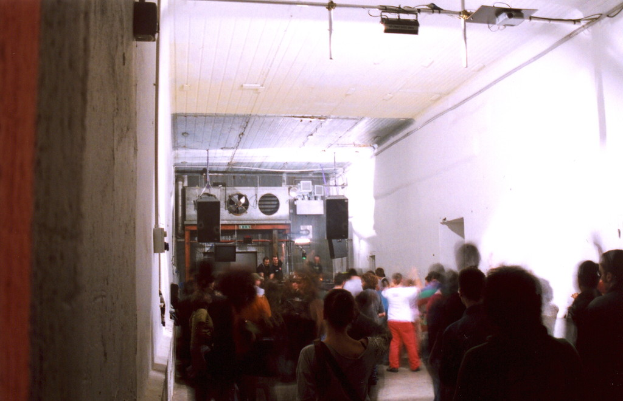 Menschen, die in der Mitte eines Raumes tanzen, mit Lautsprechern, einem Rohr und einer weißen Wand, Lichtern, einem Projektor und einem Eisenrahmen oben und Lautsprechern, einem Ventilator und anderen Gegenständen im Hintergrund.