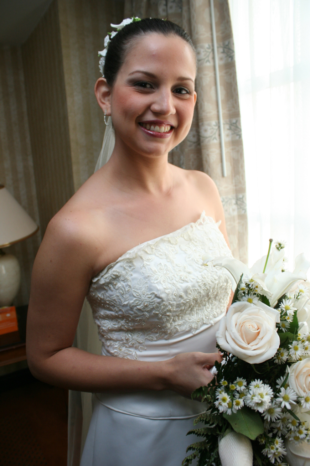 Eine strahlende Braut mit einem Blumenstrauß, vor einem Vorhang und einer Wand stehend, mit einer Lampe auf einem Tisch links.