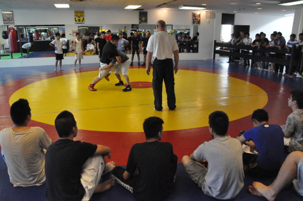 Zwei Menschen ringen auf einer Matte, daneben steht ein Mann in einem weißen T-Shirt; Zuschauer schauen von hinter einer Absperrung, einige sitzen auf dem Boden, mit Spiegeln, Boxsäcken und Postern im Hintergrund.