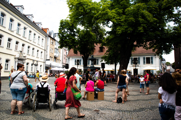 Eine Gruppe von Menschen, einige im Rollstuhl und andere stehend, geht eine Kopfsteinpflasterstraße in der Heidelberger Altstadt entlang, mit Bäumen, Gebäuden, Laternen und einer Statue im Hintergrund unter einem bewölkten Himmel.