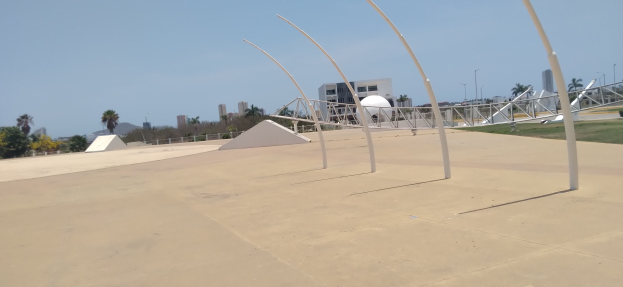 Eine Skate-Anlage mit einem zentralen Metallgebilde, umgeben von Pfählen, Gras, einem Zaun, Bäumen, Gebäuden, Straßenlaternen und einem bewölkten Himmel.