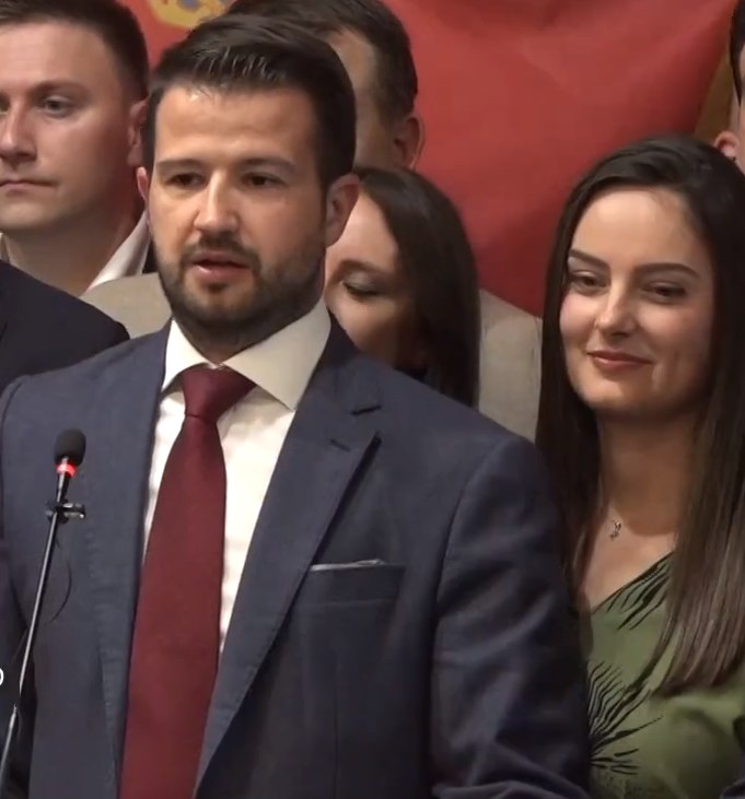 Mann im Anzug und Krawatte, der vor einem Publikum mit einem Mikrofon an einem Rednerpult steht, mit einem Banner im Hintergrund.