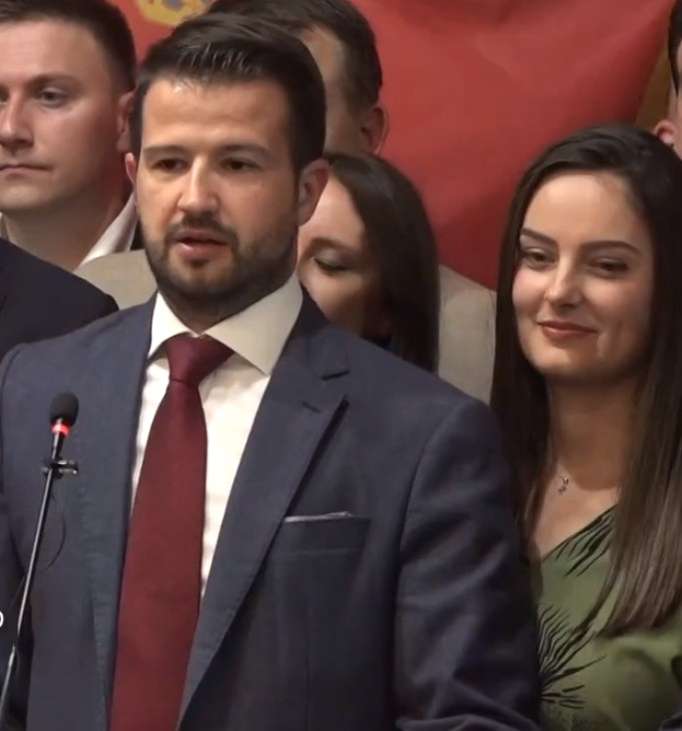 Mann im Anzug und Krawatte, der vor einem Publikum mit einem Mikrofon an einem Rednerpult steht, mit einem Banner im Hintergrund.