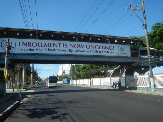 Ein Bus fährt unter einer Brücke mit einem "Jetzt Anmelden"-Schild, während Fußgänger auf dem Gehweg darunter gehen, mit Strommasten und Drähten an der Straße, Bäumen im Hintergrund und einem klaren blauen Himmel.
