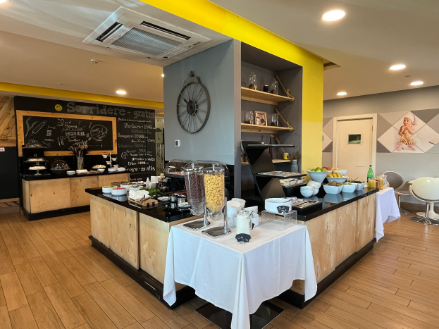 Ein Restaurantinnenraum mit Tischen und Stühlen, Lebensmitteln, einer Tafel mit Text, einer Wanduhr, Deckenleuchten, einer Klimaanlage und einer Tür im Hintergrund, gelegen im Hôtel de Ville in Paris.