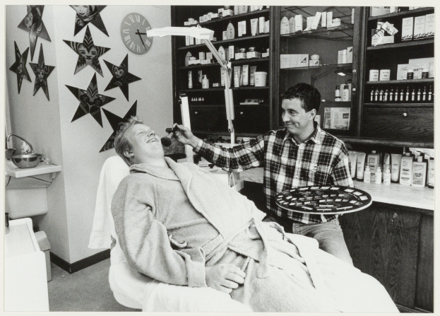 Ein Mann lächelt in einem Zahnarztstuhl, während ein anderer Mann neben ihm steht und eine Platte hält, mit einem Waschbecken, Sternen, einer Uhr und Regalen mit Flaschen im Hintergrund.