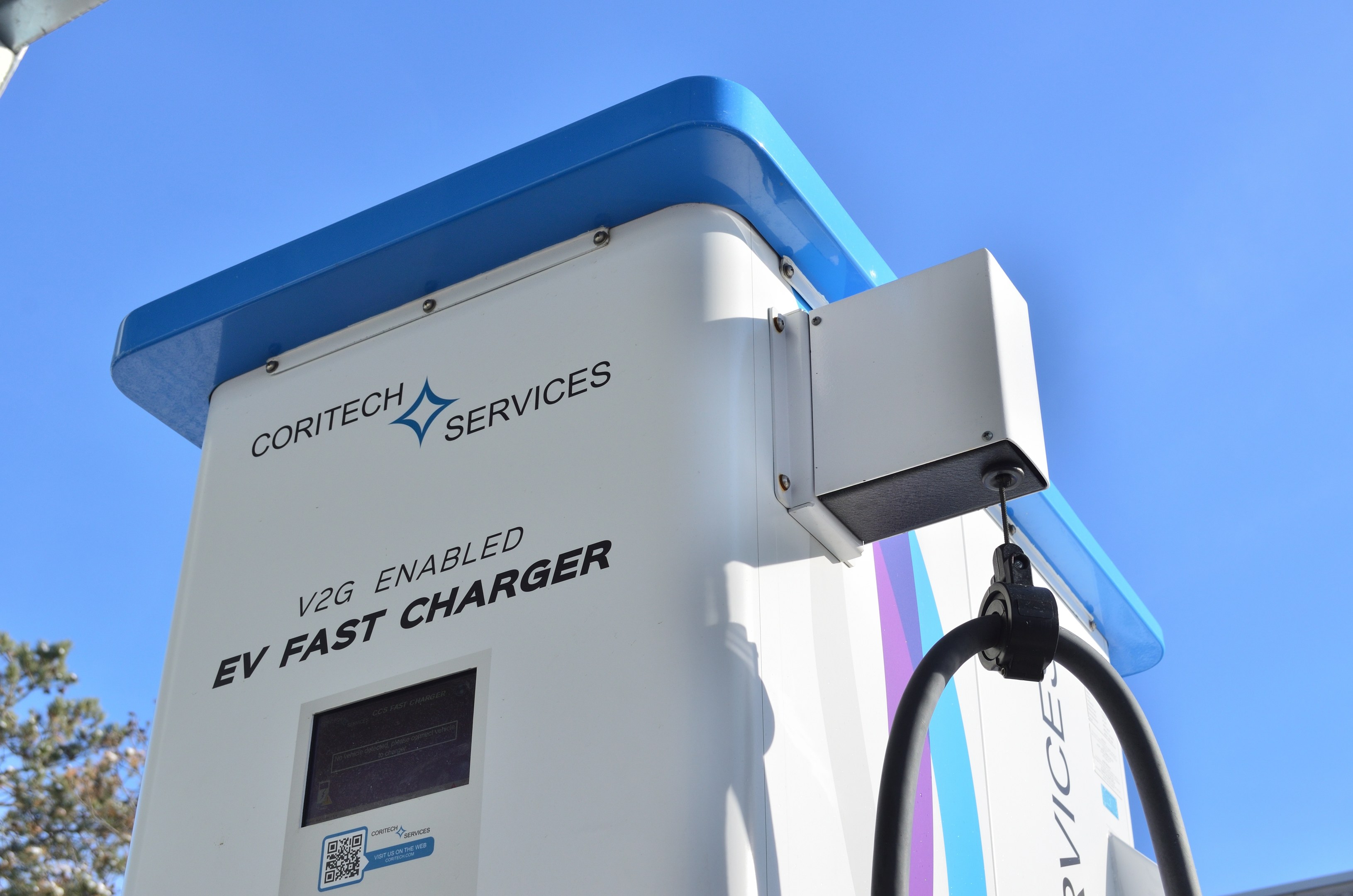 Elektrische Ladestation mit einem "V2G Fast Charger"-Schild, Bäumen, einem Gebäude und einem klaren blauen Himmel.