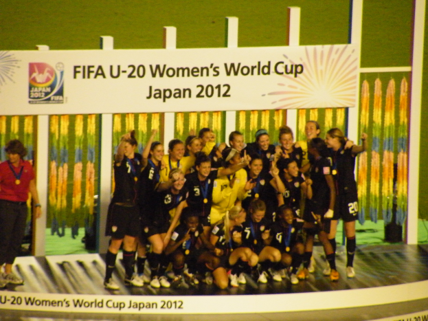 Eine Gruppe von Frauenfußballspielerinnen in schwarzen Uniformen, einige mit Medaillen, posieren auf einer Bühne mit einer Tafel im Hintergrund.
