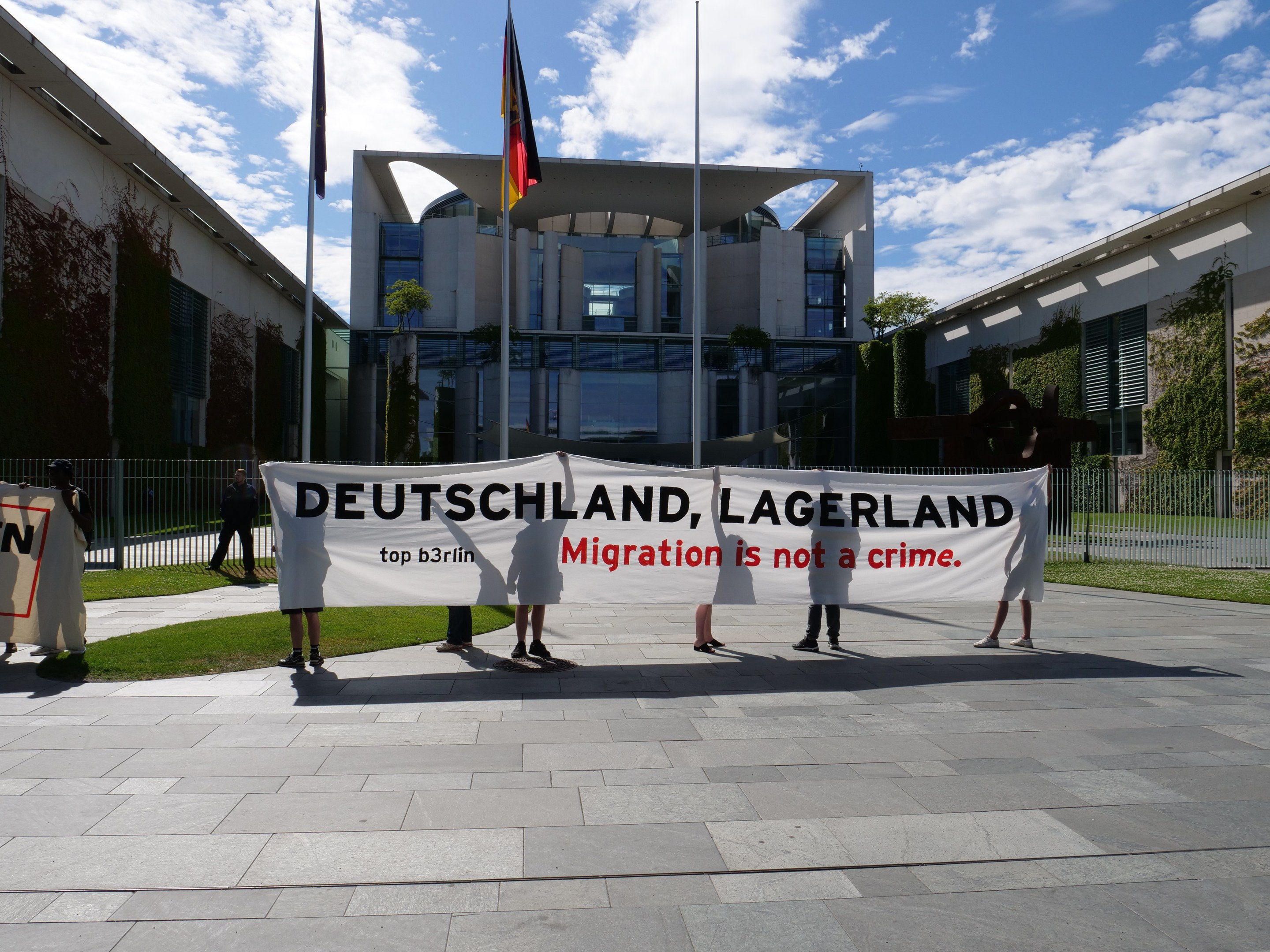Gruppe von Menschen mit einem Banner mit der Aufschrift "Deutschland, Lagerland Migration ist kein Verbrechen" vor einem Gebäude mit Fenstern, umgeben von Gras, einem Metallzaun, Pflanzen, Bäumen und Fahnenmasten vor einem bewölkten Himmel.