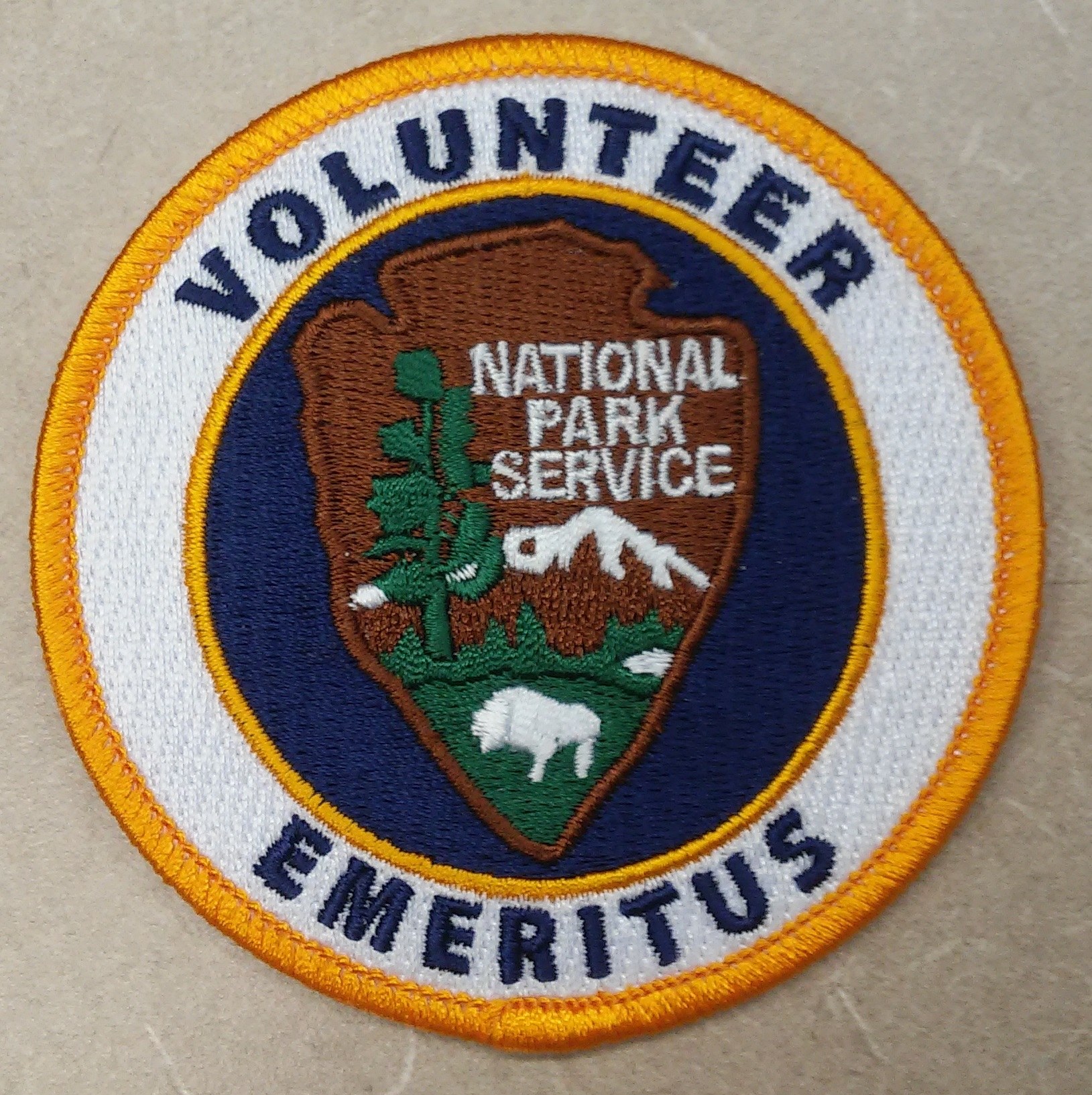 Runder Aufnäher mit "Volunteer National Park Service Emeritus" in schwarzer Schrift auf weißem Hintergrund, umrandet von einem blauen Rand und einem gelben Kreis.