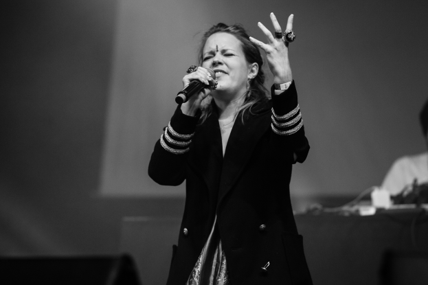 Eine Frau singt in ein Mikrofon auf einer B├╝hne, mit einem Mann, der ein Musikinstrument im Hintergrund spielt, dargestellt in Schwarz-Wei├č.