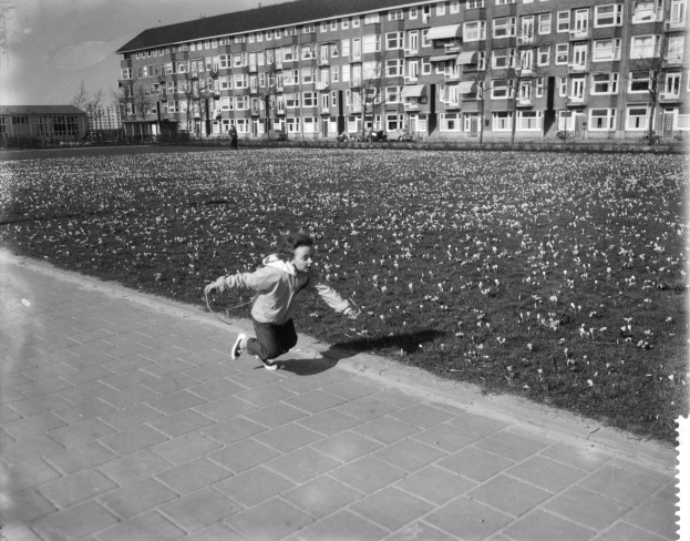 Ein junger Junge rennt über einen Gehweg in einem Park, umgeben von Gebäuden, Bäumen und einem klaren Himmel, und trägt Schuhe.