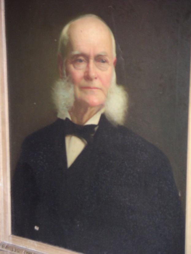 Ein gerahmtes altes Foto eines ernst aussehenden Mannes mit einem weißen Bart, Brille und ordentlich gekämmtem Haar, der schlichte Kleidung trägt.