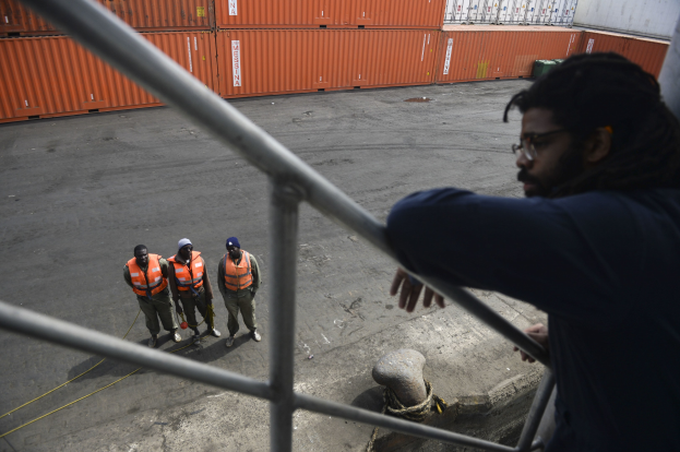 Ein Mann in legerer Kleidung steht auf einem Metallgeländer neben einer Gruppe von Menschen in orangen Jacken und Mützen, mit Containern und einer Wand im Hintergrund am Hafen von Los Angeles.